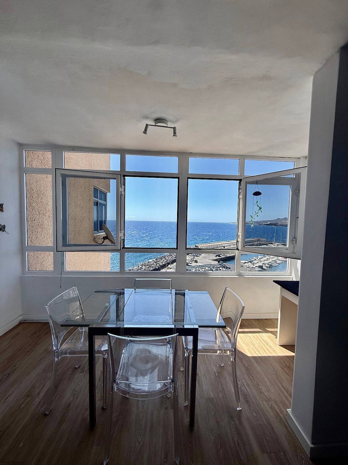 Glass dining table with four clear chairs in front of a wide window onto the sea and the harbour