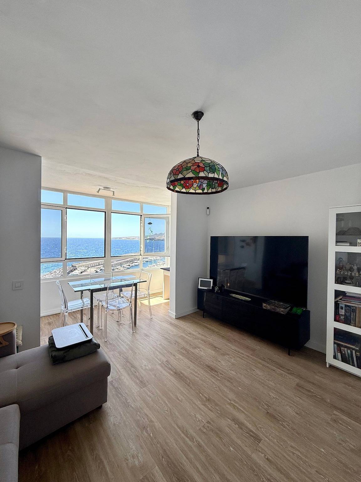 Living room with sofa, television and a stained-glass pendant lamp, the window and the sea behind